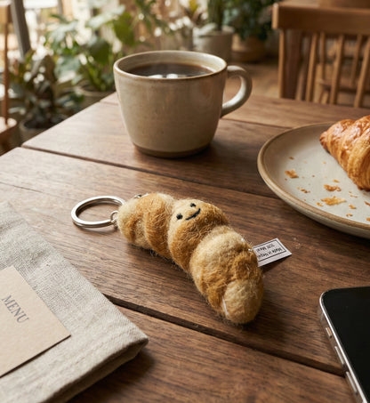 Golden brown felt croissant keychain on wooden cafe table next to coffee and real croissant