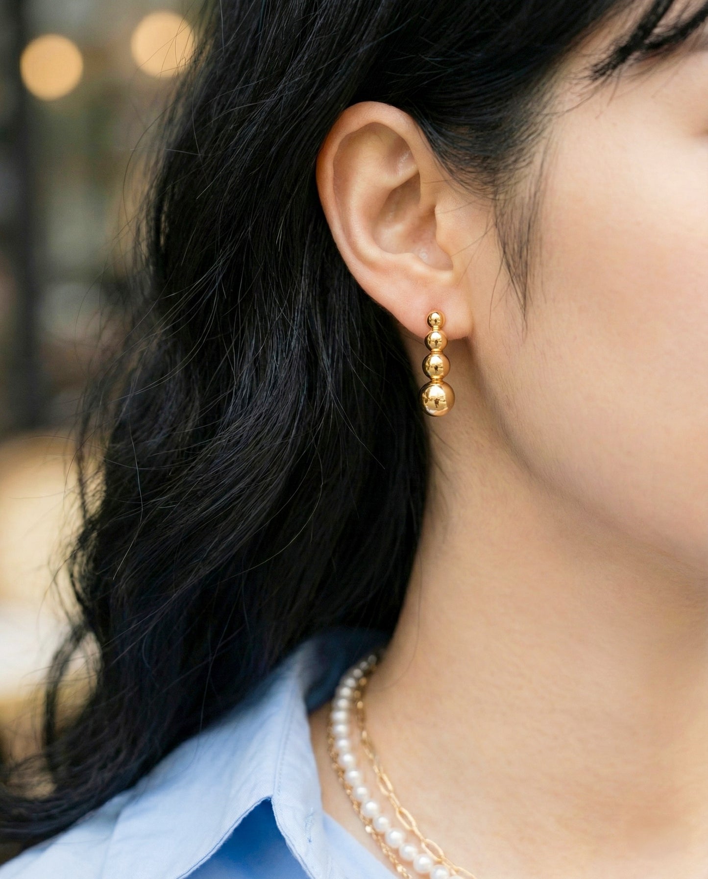 Gold graduated sphere bar earrings styled with blue blouse and pearl necklace layers