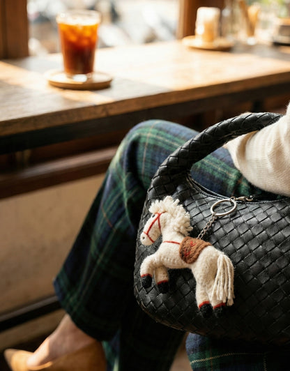 Wool felt horse bag charm hanging on black leather woven shoulder bag with plaid skirt