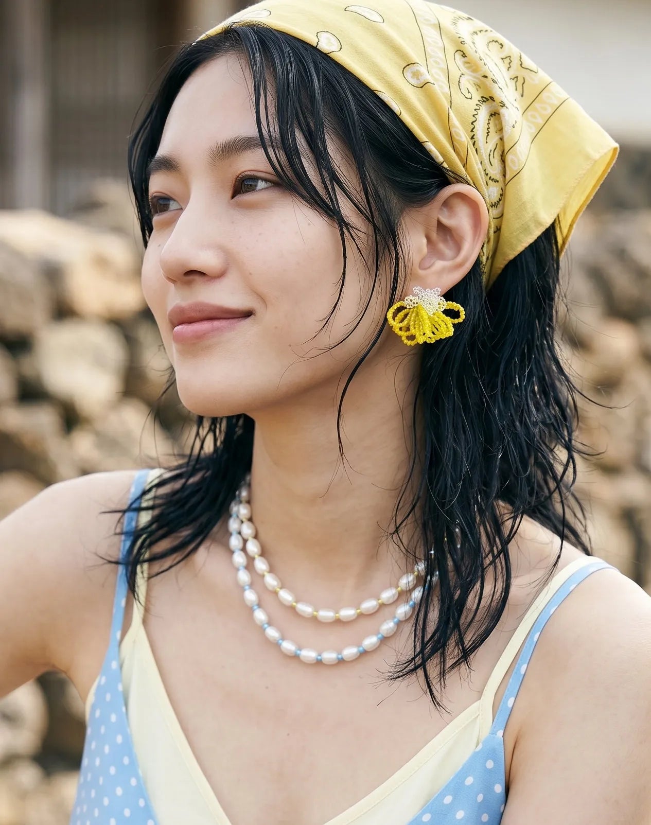 A lifestyle shot of a woman with trendy wet-look hair tucking a strand behind her ear. She models vibrant yellow beaded earrings paired with layered blue and black bead necklaces and a purple dress.