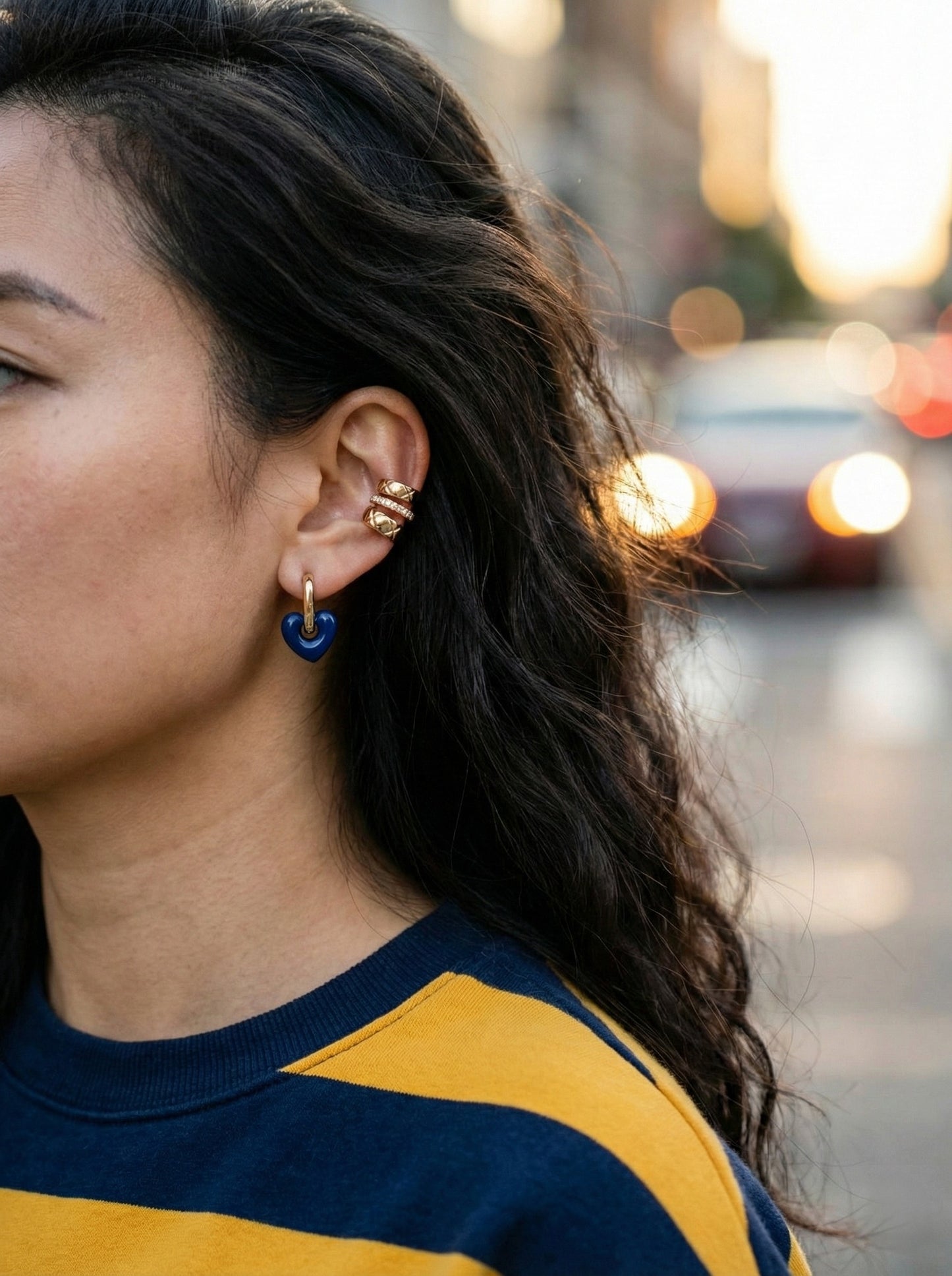 Woman wearing navy blue heart huggie earrings with yellow and blue colorblock sweater, side ear view