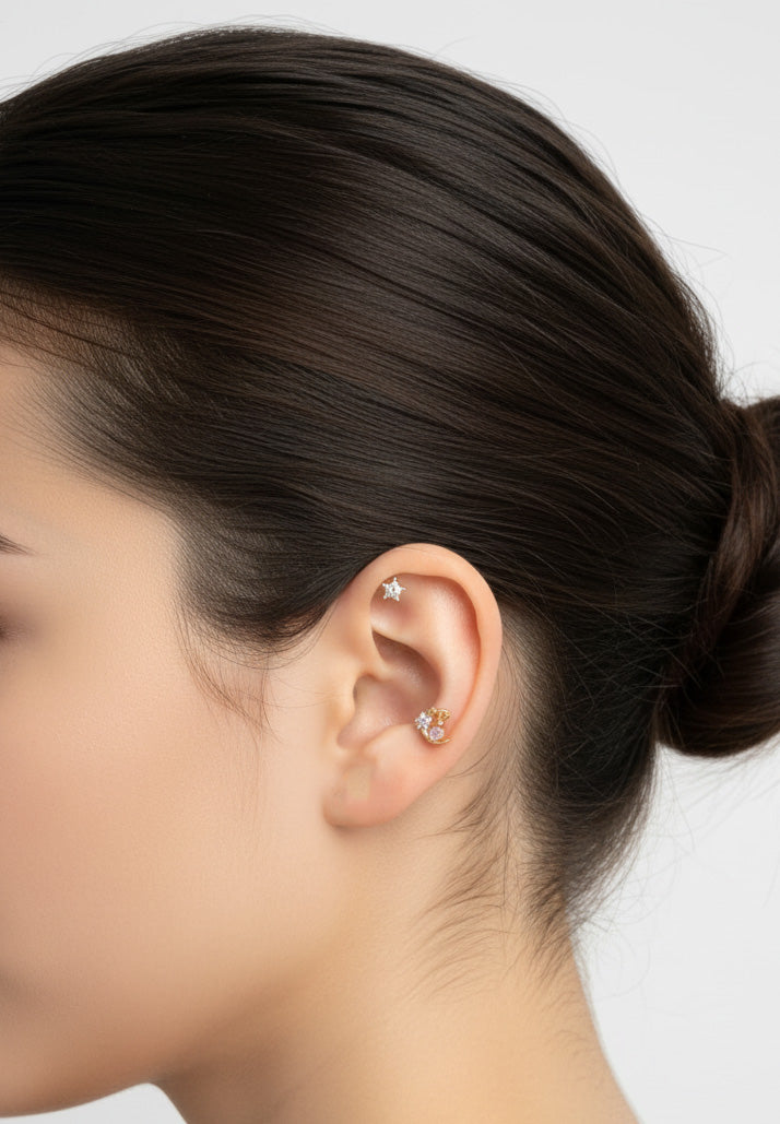 Elegant left profile of a woman’s ear wearing a star cartilage stud and a gold moon earring on the conch, with hair tied in a low bun and neutral backdrop.