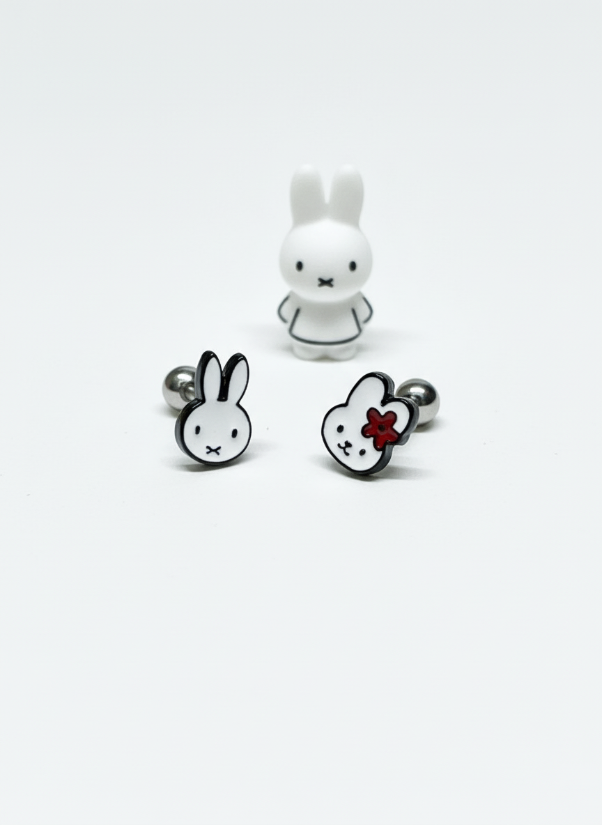 Minimalist white background with Miffy barbell earrings placed in front of a monochrome Miffy figurine; one earring features Miffy with a red flower, the other a plain version.