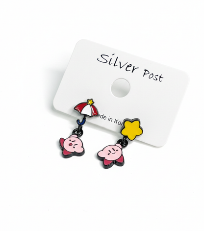 Close-up of mismatched Kirby earrings on a display card — one with red umbrella and flying Kirby, the other with a yellow star and smiling Kirby.
