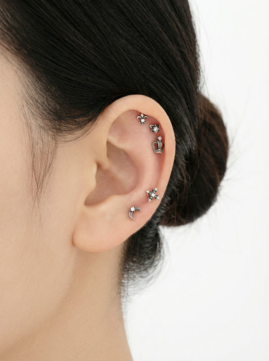 close-up of a woman’s ear styled with gothic barbell studs in star, moon, cross, fleur-de-lis, and crown shapes, silver surgical steel, multiple cartilage piercings