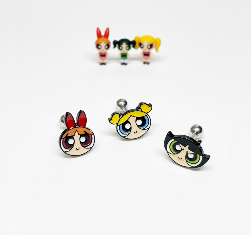 three Powerpuff Girls–inspired character barbell earrings arranged in front with matching mini figures blurred in the background