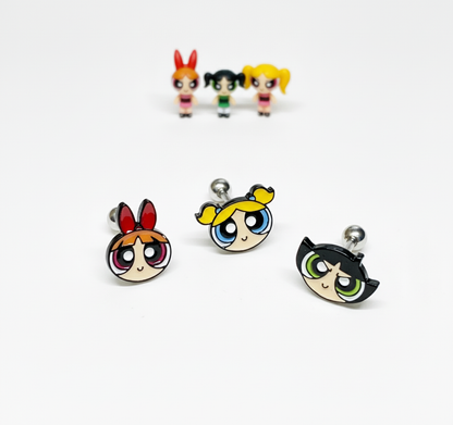 three Powerpuff Girls–inspired character barbell earrings arranged in front with matching mini figures blurred in the background