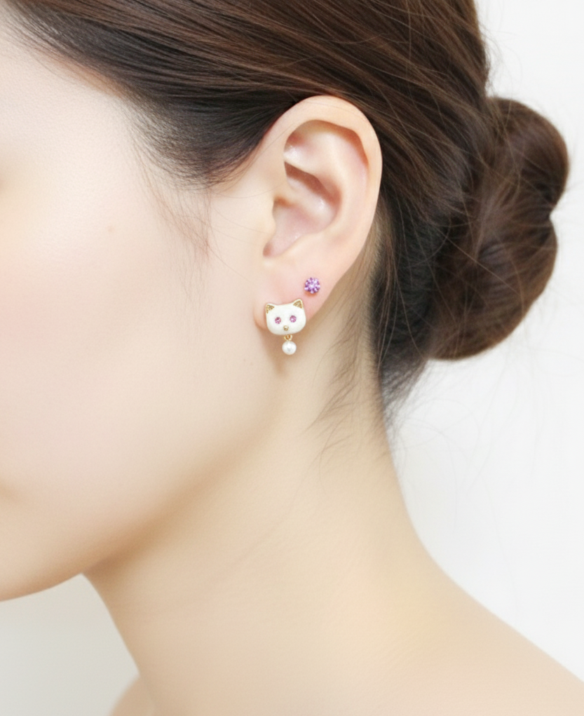 Model wearing white enamel cat stud earrings with a tiny pearl drop, paired with a small lavender stud on the upper lobe