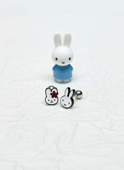 Two stainless steel barbell earrings featuring Miffy characters — one with a red flower, the other with simple ears — displayed with a small Miffy figurine in a blue dress on crumpled white paper.