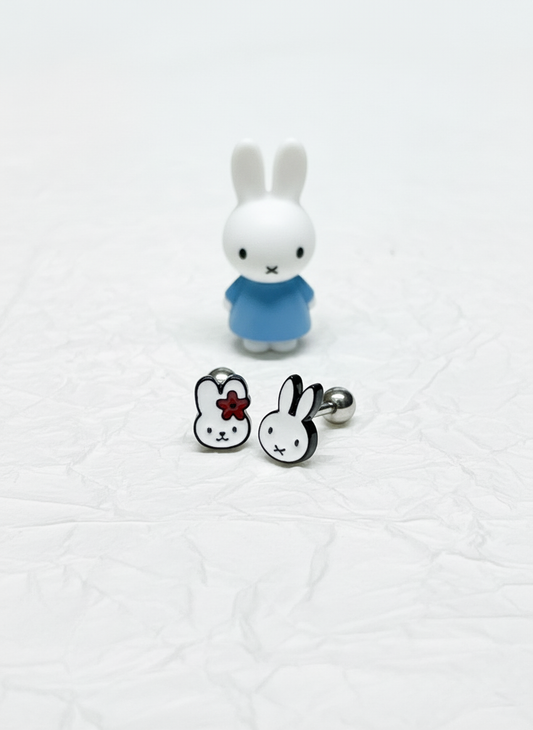 Two stainless steel barbell earrings featuring Miffy characters — one with a red flower, the other with simple ears — displayed with a small Miffy figurine in a blue dress on crumpled white paper.