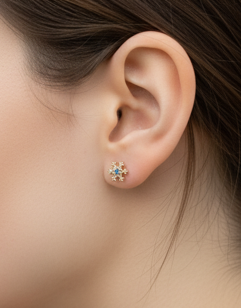 Gold snowflake stud earring with blue center stone, worn on earlobe, styled against natural skin tone.