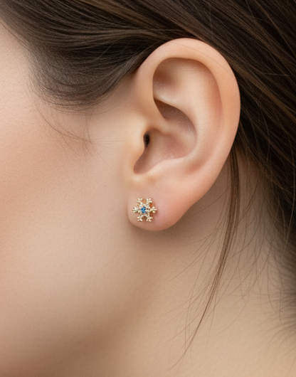 Gold snowflake stud earring with blue center stone, worn on earlobe, styled against natural skin tone.