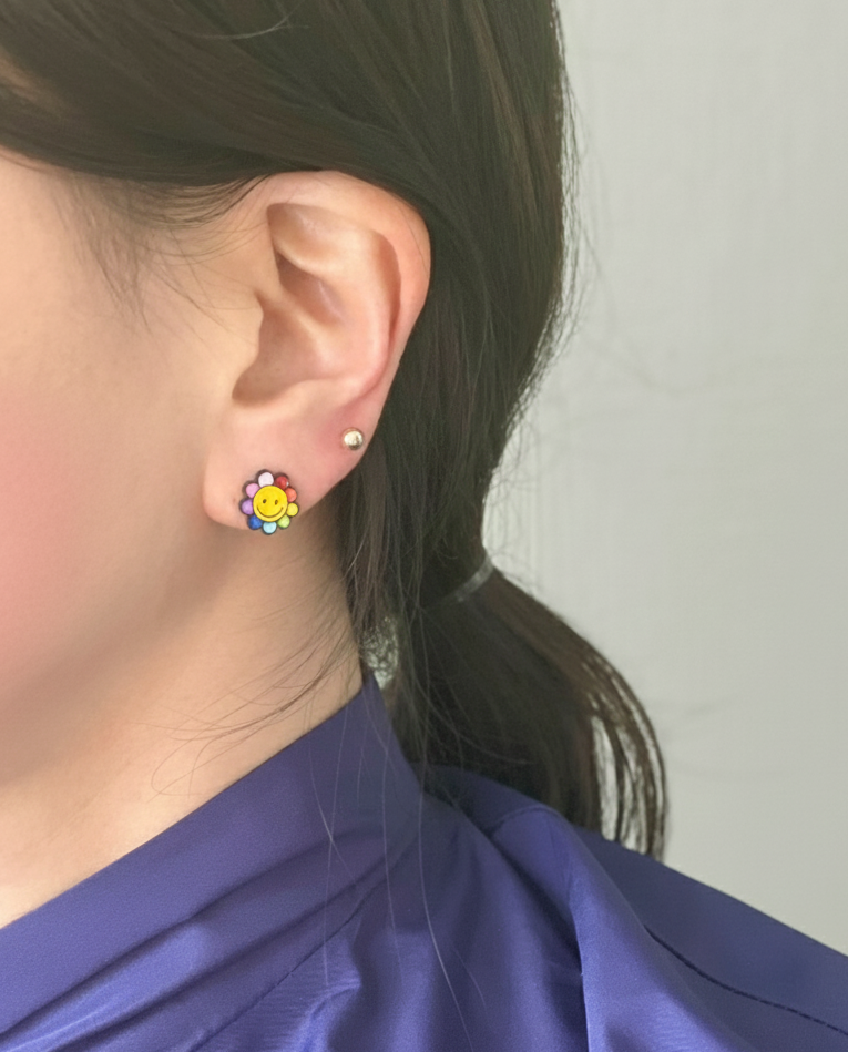 Colorful smiley flower earring with rainbow petals worn on ear, paired with a gold stud above