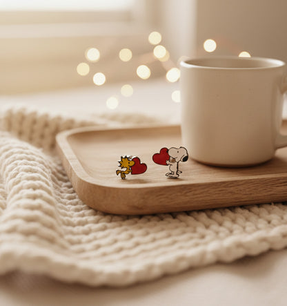 Snoopy and Woodstock heart barbell earrings styled on a wooden tray next to a white cup with cozy lighting.