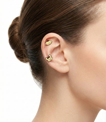 Model's ear wearing two Pompompurin character barbell piercings — one smiling and one neutral — styled in inner conch and helix placements.
