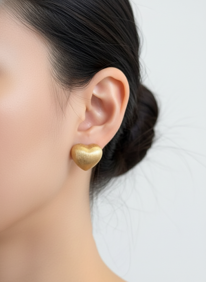 gold brushed-metal heart stud worn on the ear, showing its rounded volume and soft matte glow
