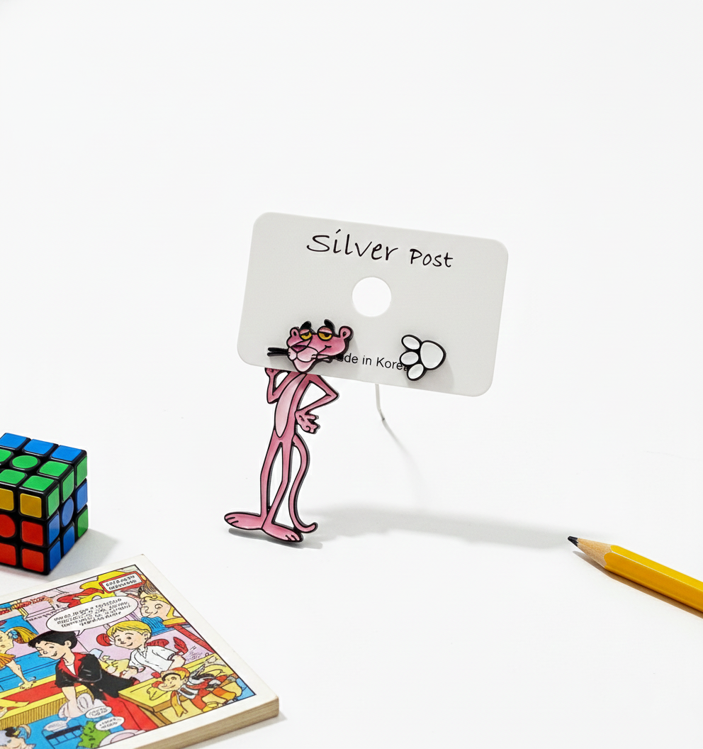 Pink Panther mismatched earrings displayed on a white earring card, with a Rubik's cube, pencil, and comic book in the background.