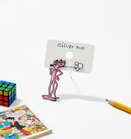 Pink Panther mismatched earrings displayed on a white earring card, with a Rubik's cube, pencil, and comic book in the background.