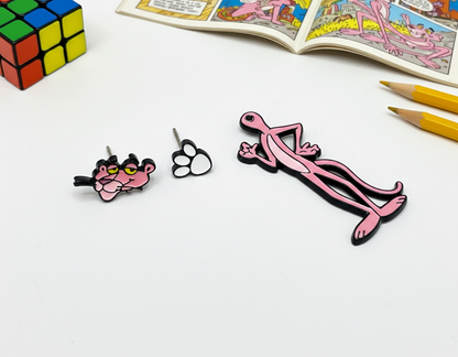 Close-up of Pink Panther head and paw stud earrings laid flat, with the Panther body charm next to them, comic book and pencils in the background.