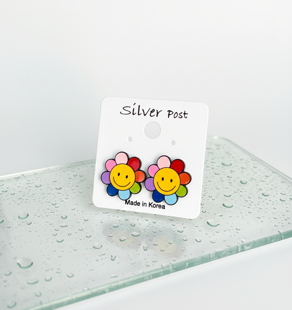 Colorful smiley flower stud earrings with rainbow petals displayed on a white earring card labeled "Silver Post" with "Made in Korea," set against a glass surface with water droplets.