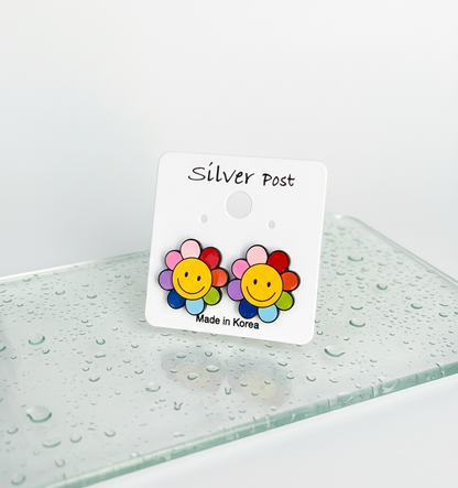 Colorful smiley flower stud earrings with rainbow petals displayed on a white earring card labeled "Silver Post" with "Made in Korea," set against a glass surface with water droplets.