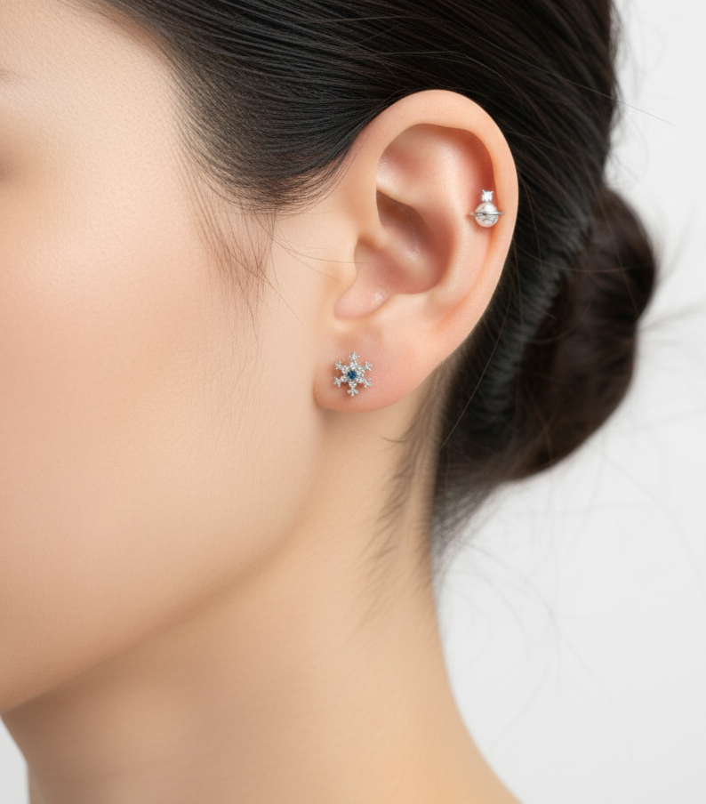 Blue snowflake stud earring on earlobe, paired with a planet-shaped stud on upper cartilage.