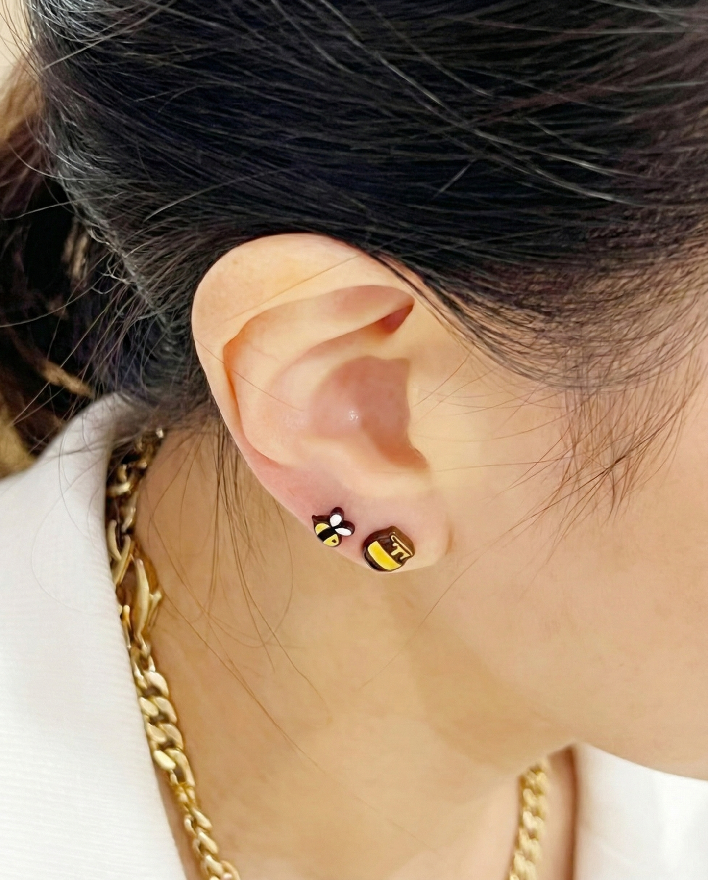 Close-up of ear wearing bee and honey pot earrings side by side, styled with gold chain necklace.
