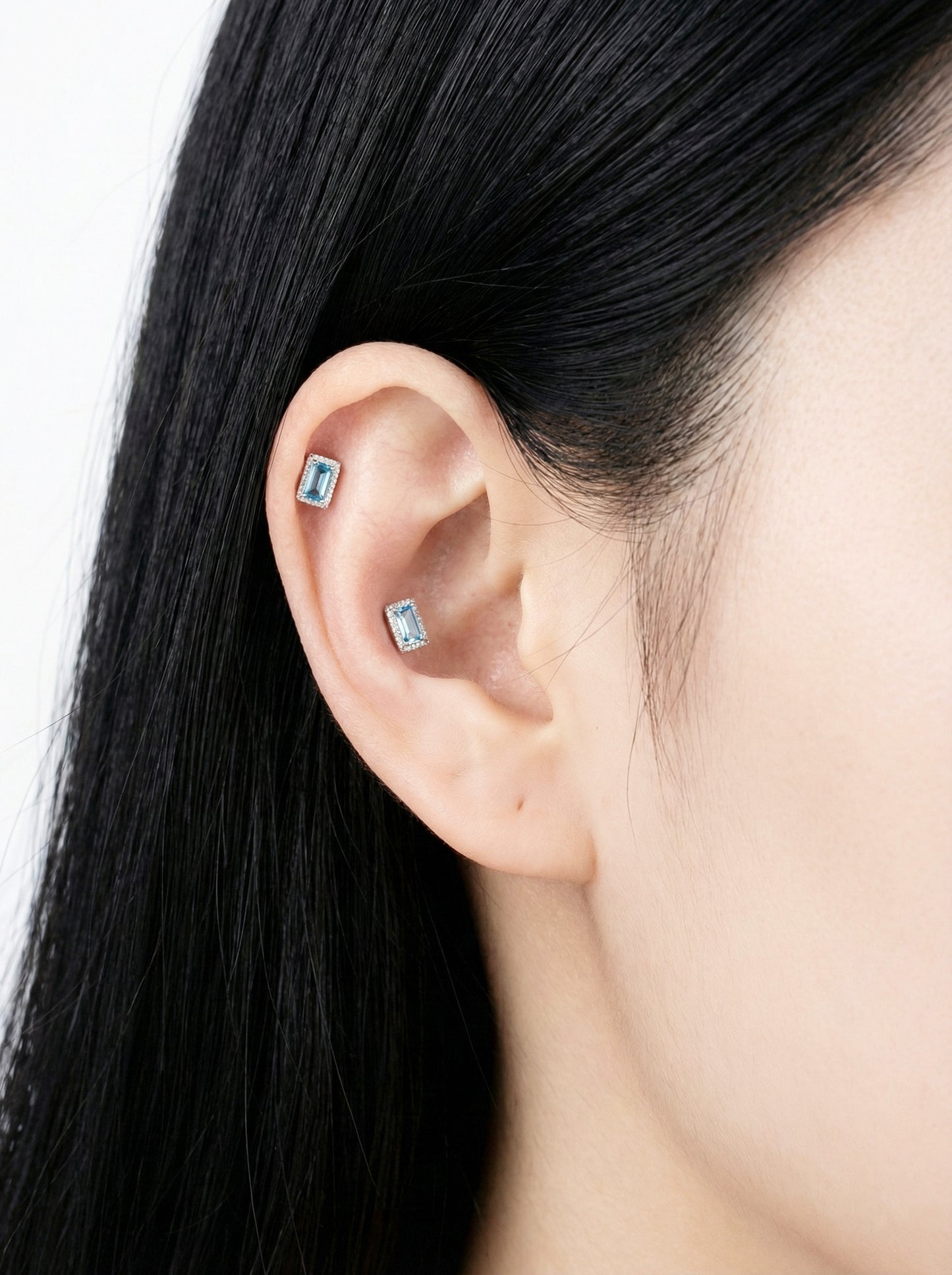 Close-up of ear wearing two rectangular blue CZ barbell earrings in helix and conch positions.