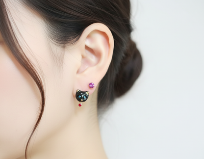 Model wearing black enamel cat stud earrings with a red crystal drop, paired with a small lavender stud above