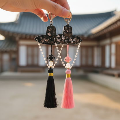 Hand holding a pair of Korean-style gat tassel keychains with mother-of-pearl and pearl detailing in front of a traditional house