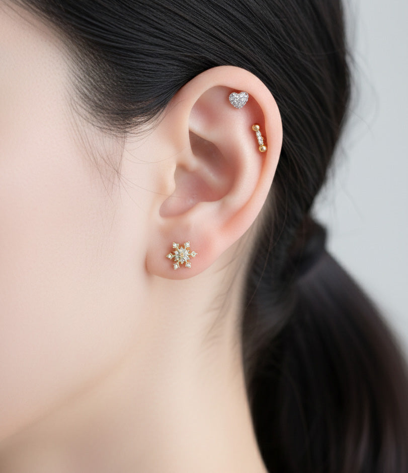 Model wearing a snowflake earring on the lobe, silver heart stud on helix, and gold barbell on rook.