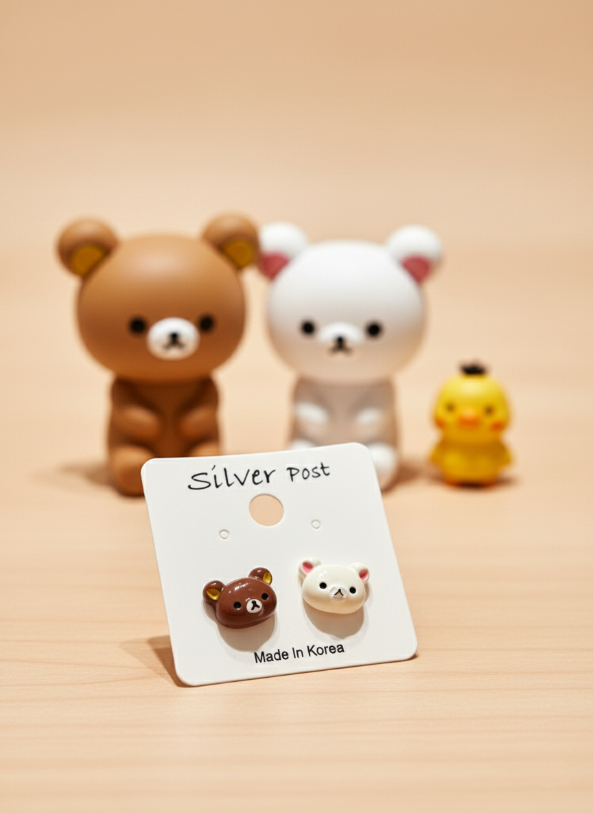 Brown and ivory bear-head stud earrings displayed on a white Silver Post card, with blurred Rilakkuma-style bear figurines in the background.
