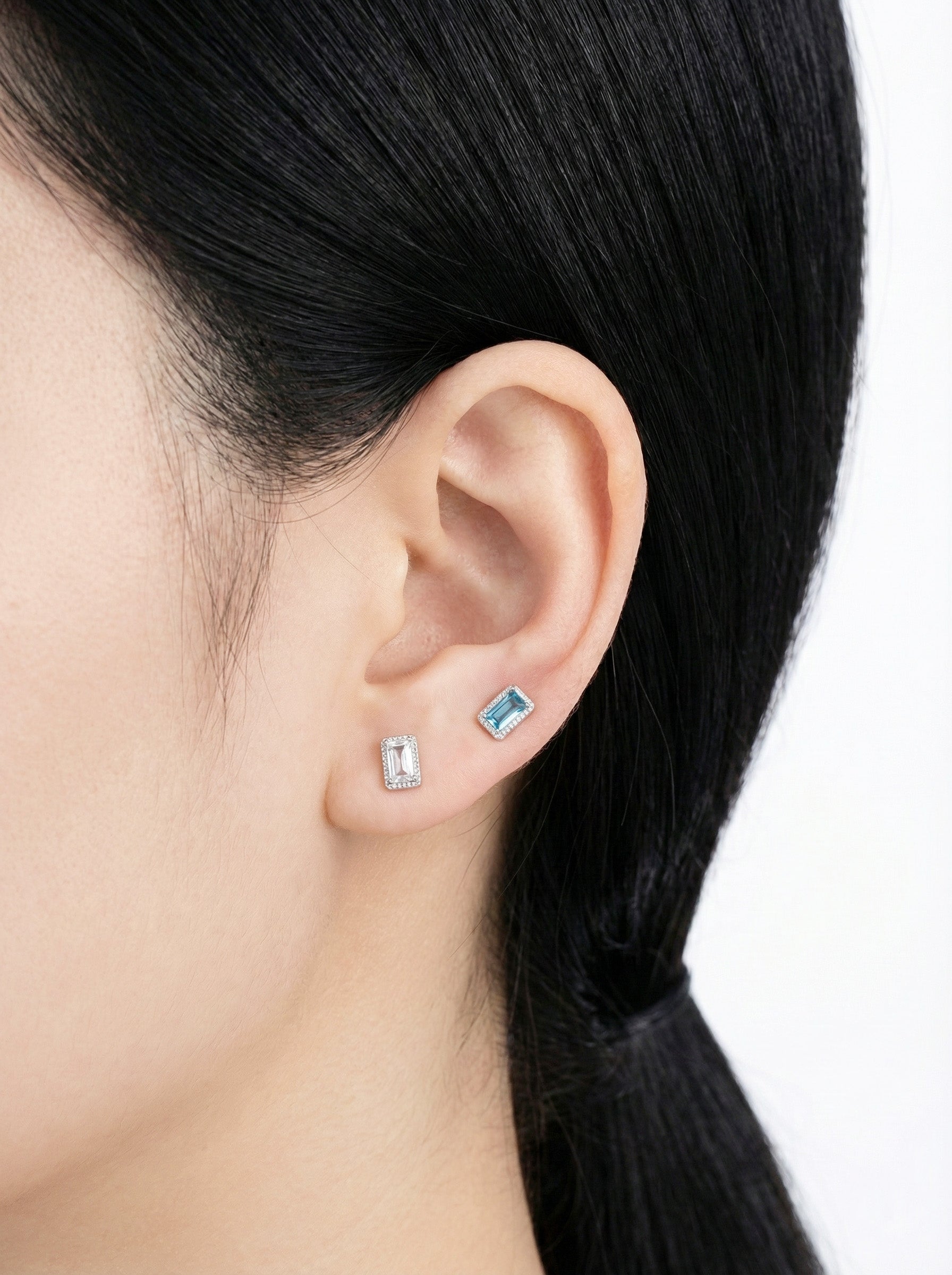 Close-up of ear wearing one rectangular blue and one clear CZ barbell earring stacked on earlobe.
