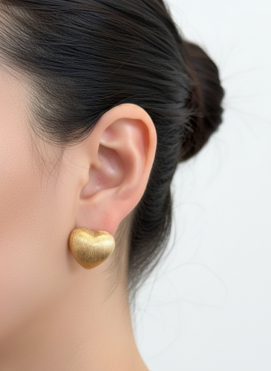 gold brushed-metal heart stud earring on the ear, highlighting the smooth matte finish and domed heart shape