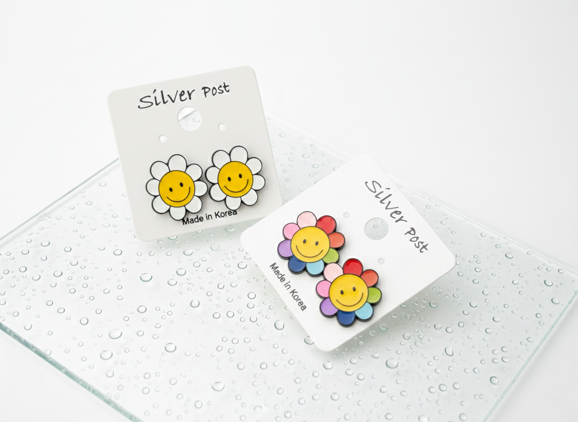 Two pairs of smiley flower earrings, one white petal and one rainbow petal, each on their own card, placed diagonally on a misted glass surface.
