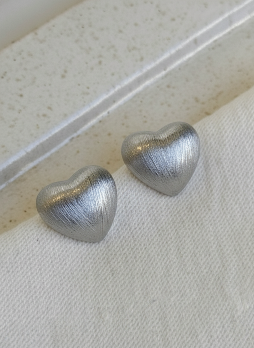 silver brushed-metal heart stud earrings placed on a folded cream fabric with a textured stone background.