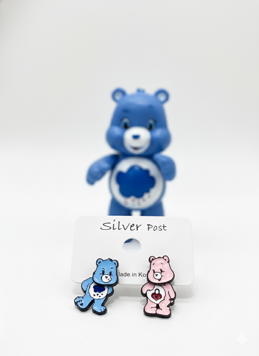 Focused front shot of pink and blue bear earrings on a white card, with a large blue bear toy softly blurred in the background.