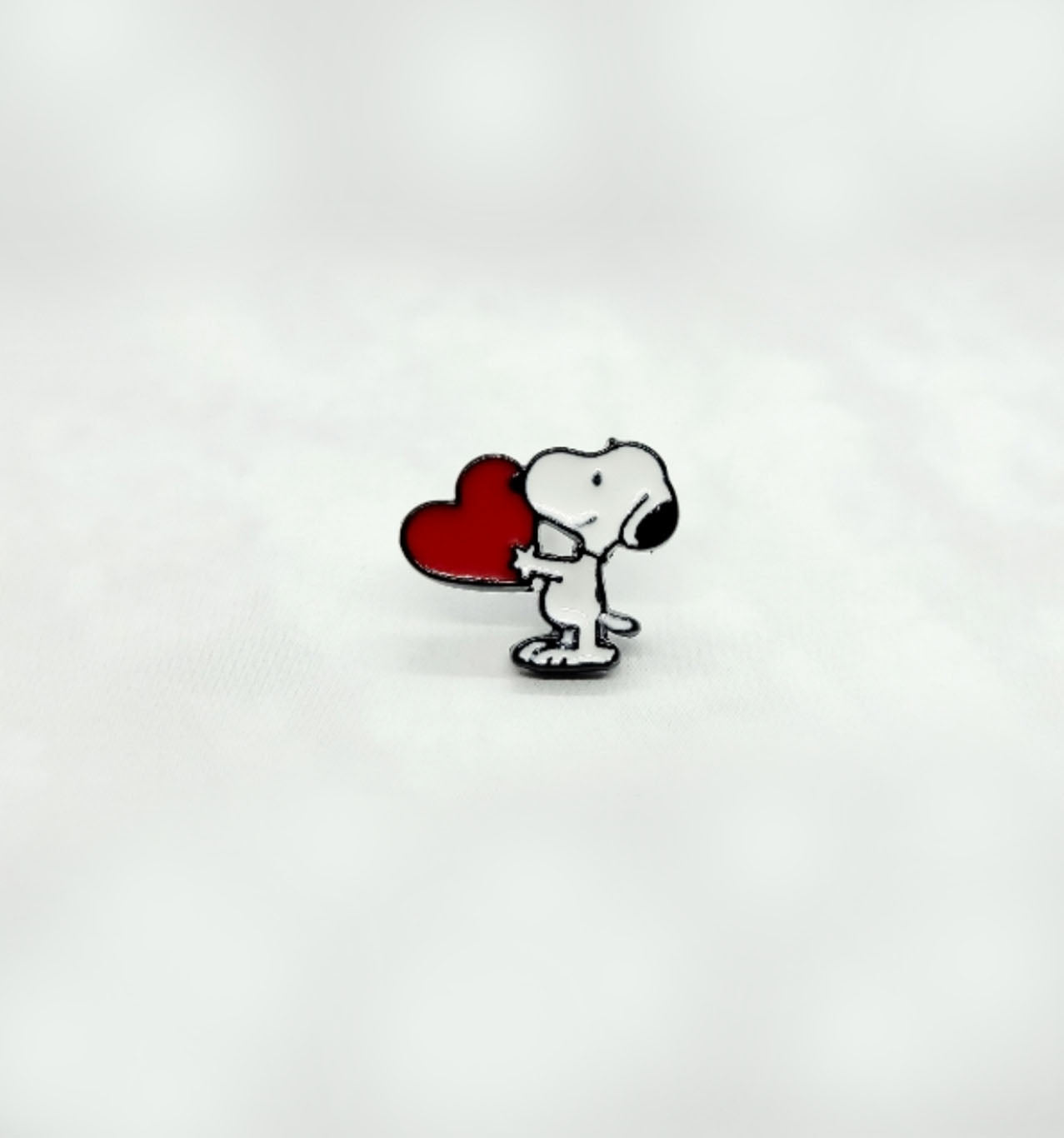 Solo Snoopy enamel earring holding a red heart, photographed upright on white fabric.