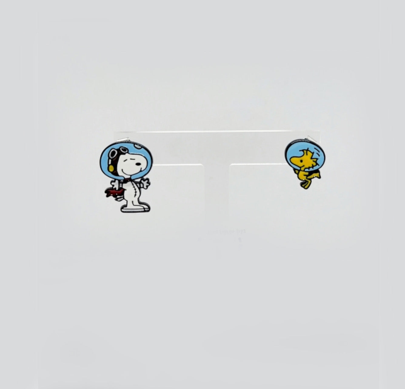 Cartoon astronaut Snoopy and Woodstock enamel earrings displayed on a translucent white stand.