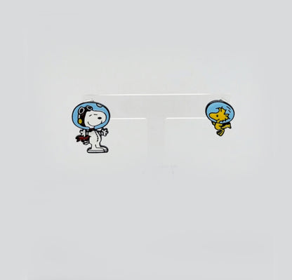 Cartoon astronaut Snoopy and Woodstock enamel earrings displayed on a translucent white stand.
