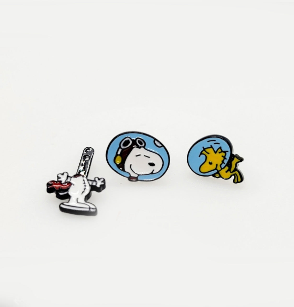 Three enamel earring designs on a white background: Snoopy with rocket, Snoopy in a helmet, and Woodstock in a helmet.
