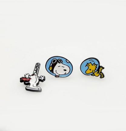 Three enamel earring designs on a white background: Snoopy with rocket, Snoopy in a helmet, and Woodstock in a helmet.