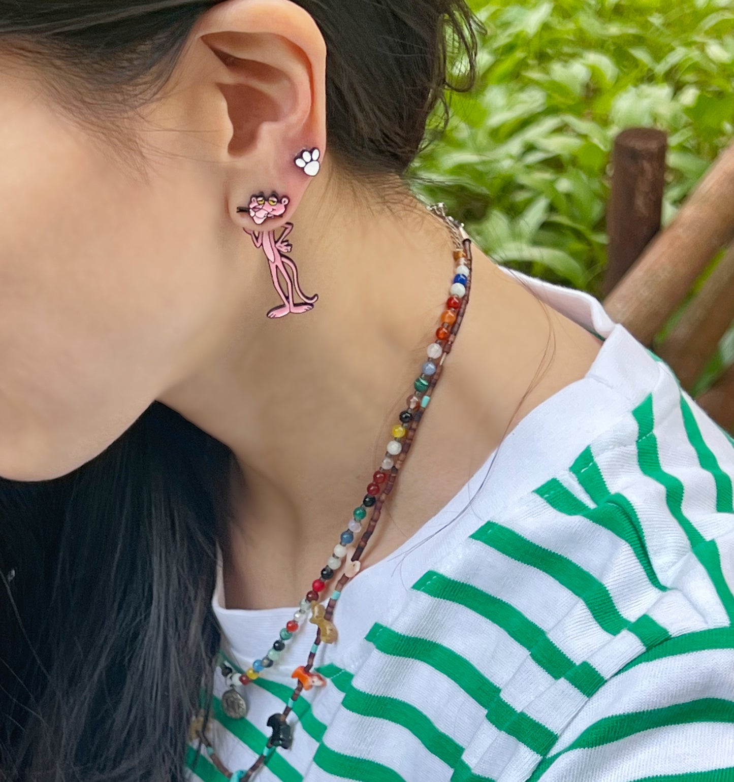 Model wearing mismatched Pink Panther charm and paw stud earrings outdoors, paired with a multicolor beaded necklace and green striped shirt.