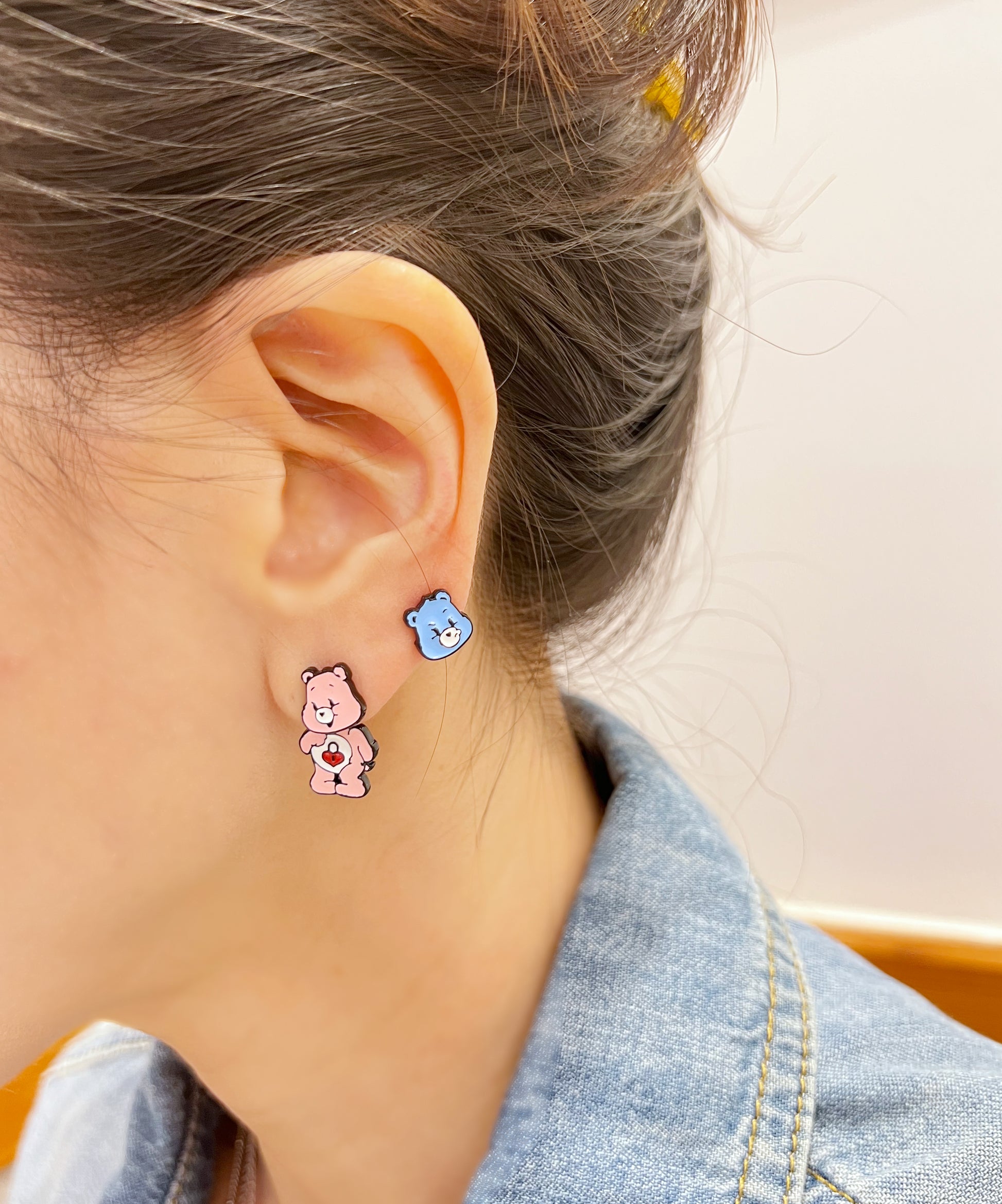 Side view of pink and blue bear earrings styled on opposite ears, worn with short hair tucked behind and a soft smile.