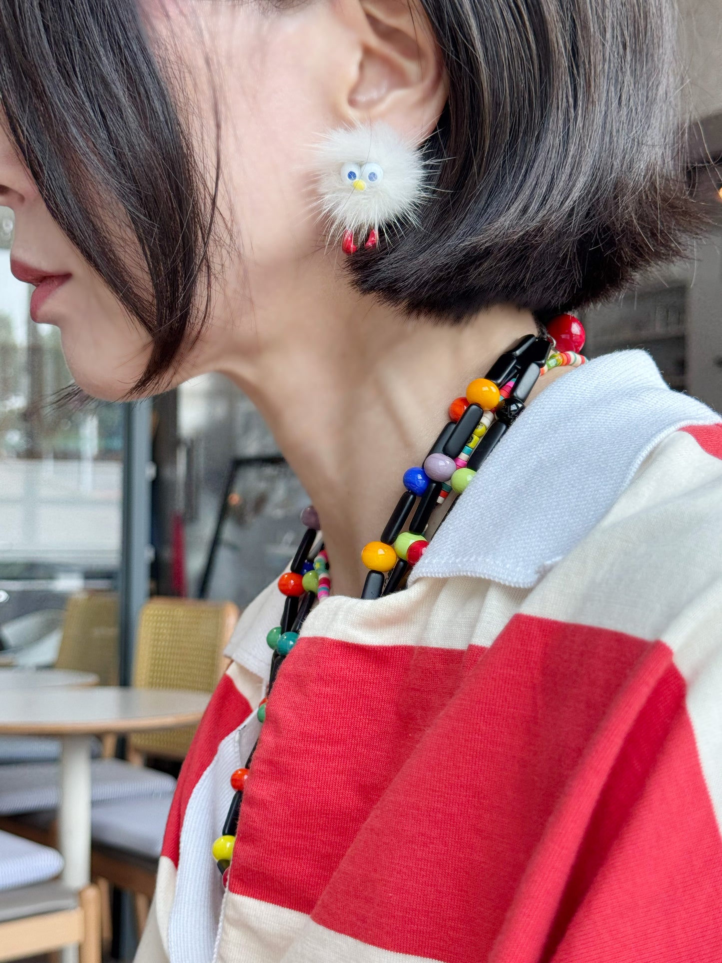 Person wearing a white fluffy earring with red feet, styled with colorful beaded necklace