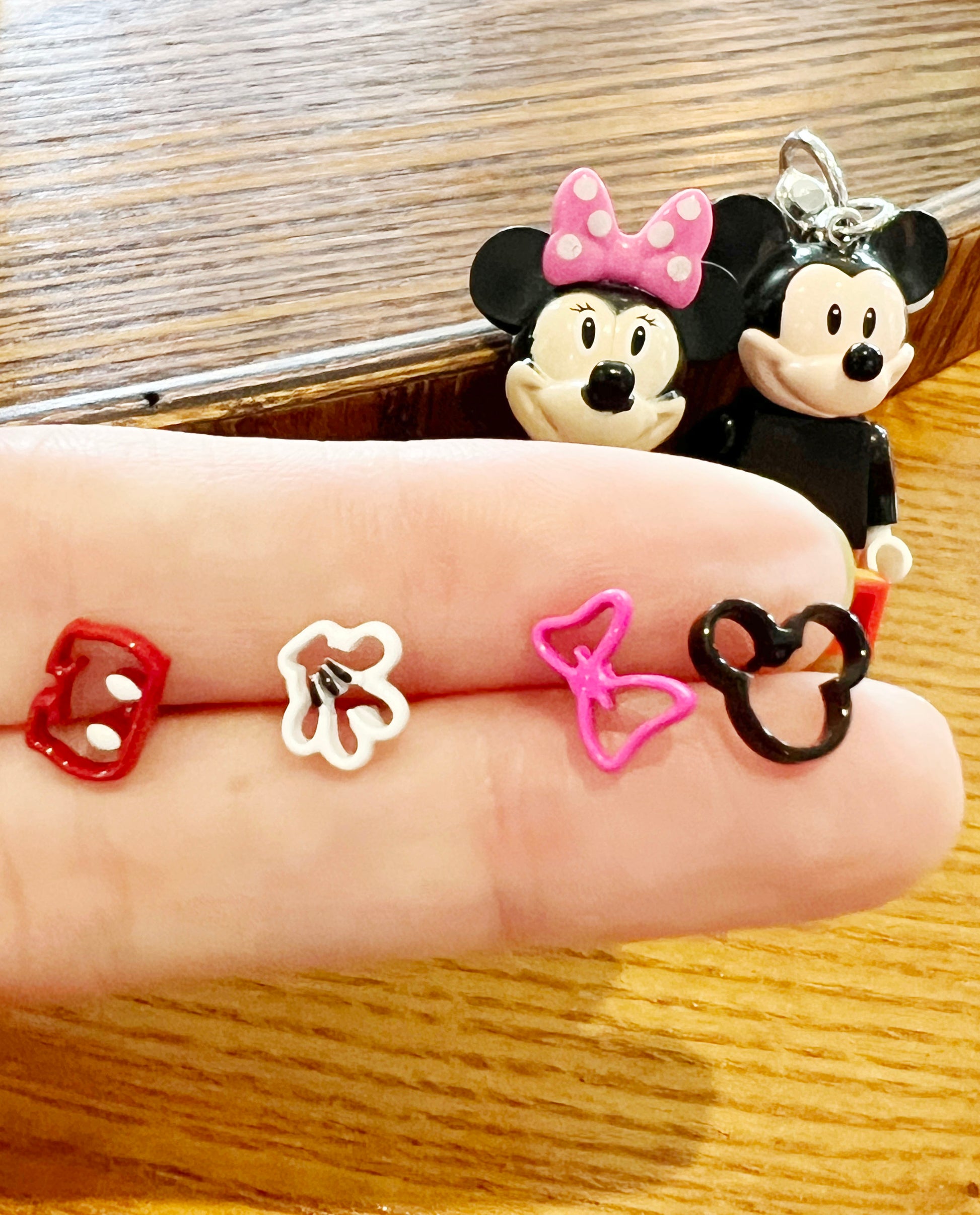 Mickey character earrings displayed across a fingertip — red pants, white glove, pink bow, and black head — with Minnie and Mickey keychains in the background.
