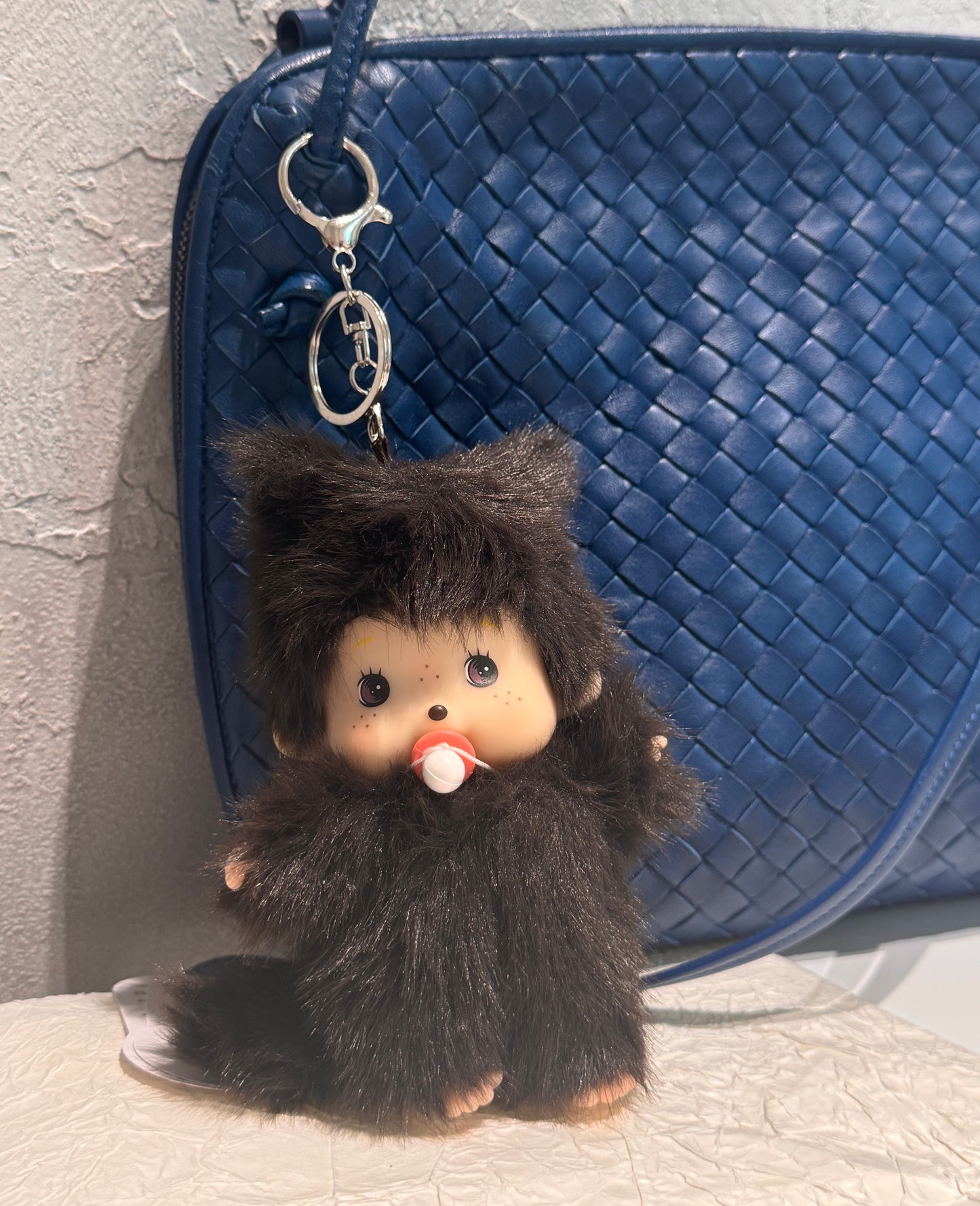 Plush doll keychain hanging from a blue woven leather handbag, set against a light concrete wall