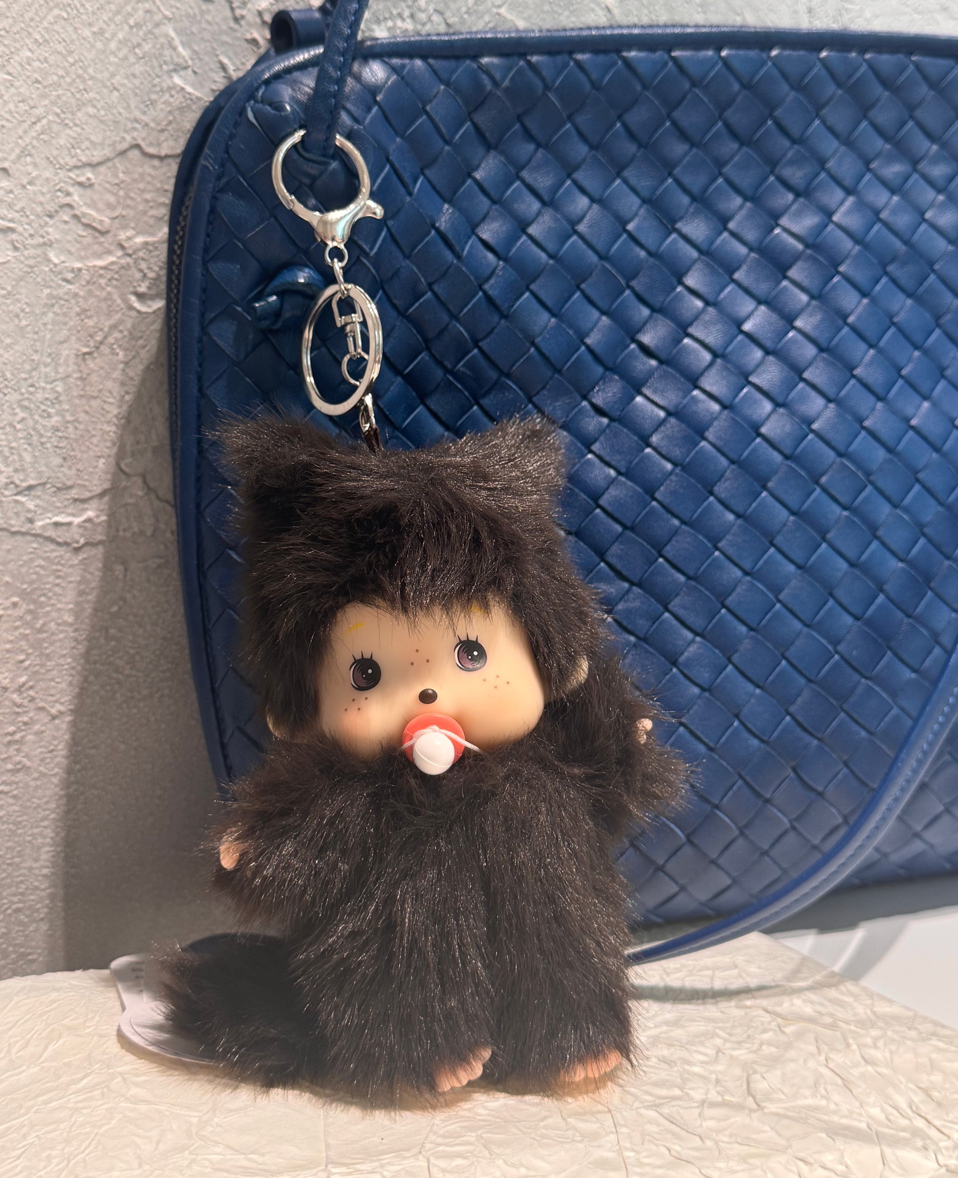 Plush doll keychain hanging from a blue woven leather handbag, set against a light concrete wall