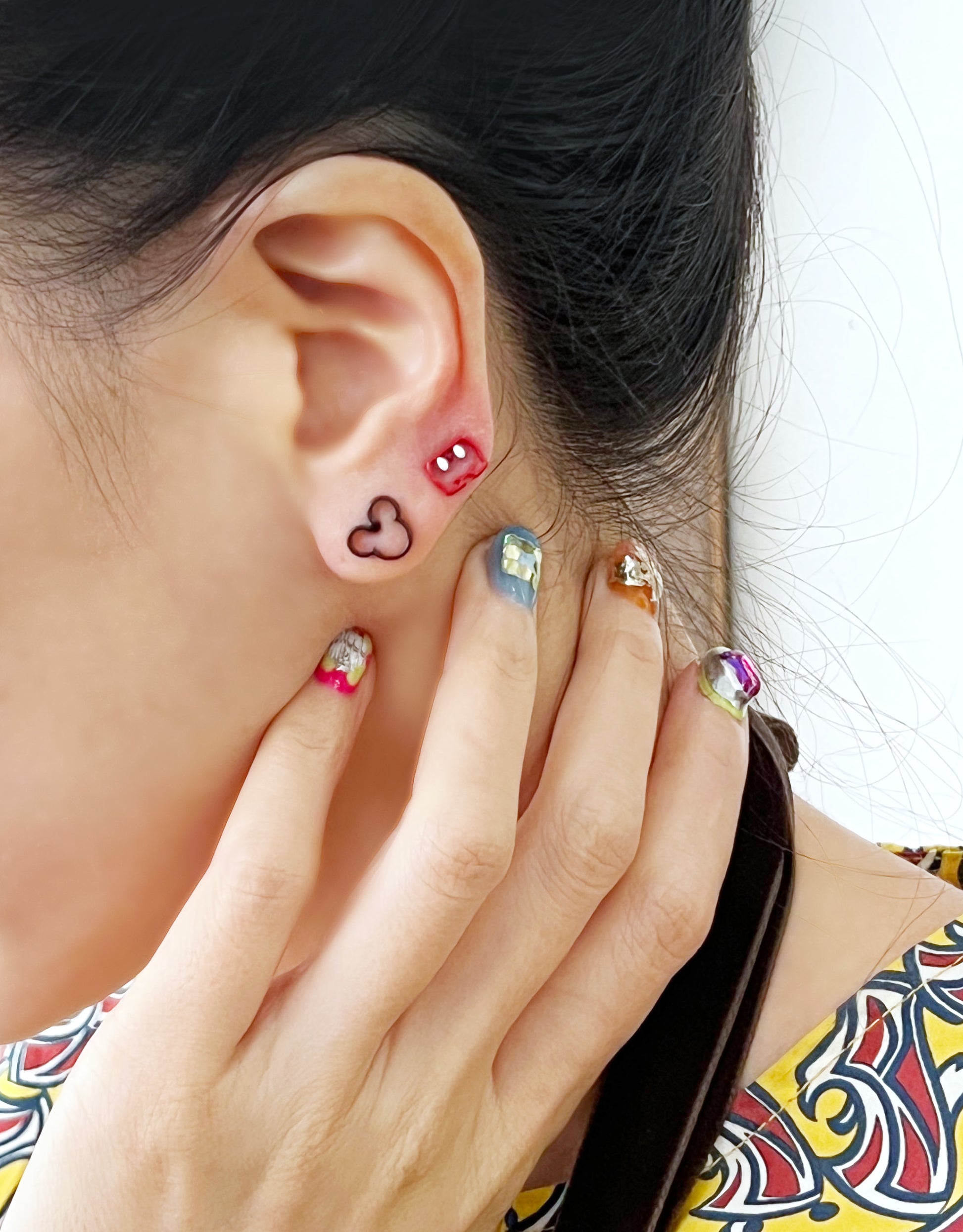 Close-up of ear wearing red shorts and black Mickey silhouette earrings together, styled with bold patterned top and vibrant multicolor nails.