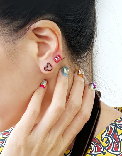 Close-up of ear wearing red shorts and black Mickey silhouette earrings together, styled with bold patterned top and vibrant multicolor nails.
